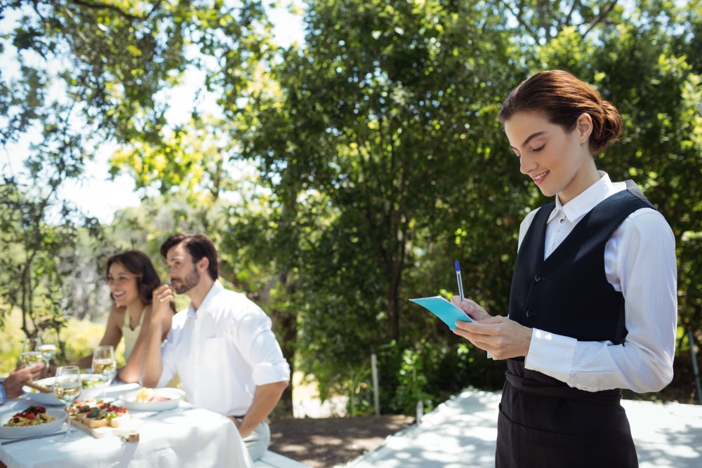 Mujer vestida como una camarera tomando notas en una libreta para seguir organizando una boda, de fondo una familia disfruta de una comida
